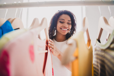 Joyful young woman browsing clothes rack in living room during daylight for shopping or donationの写真素材