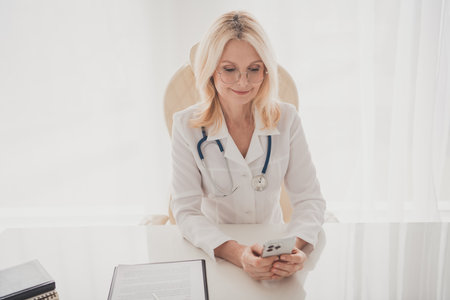 Senior female doctor in medical office using smartphone with a smile, emphasizing modern healthcare communication and prescription guidanceの写真素材