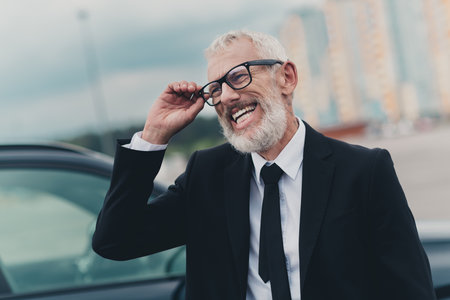 Senior businessman smiling confidently outside in parking area next to a carの写真素材