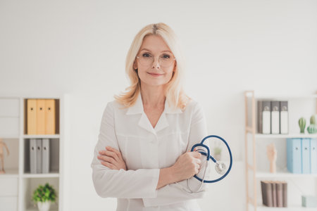 Confident mature female doctor in a clinical office holding stethoscope, embracing healthcare profession with compassionの写真素材