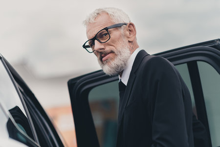 Confident senior businessman stepping out of car, wearing formal suit, on city street in an urban...