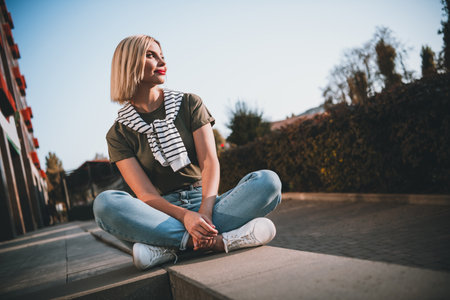 Full body photo of young blonde woman sit concrete border wondered wear khaki clothes tied shoulders sweater walk outside urban city parkの写真素材