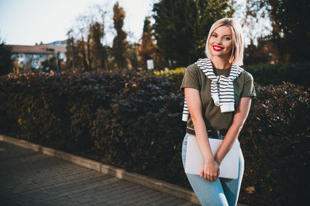 Photo of positive young blonde girl hold netbook student wear khaki outfit tied shoulders sweater walk park sunny spring weather outsideの写真素材