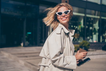 Photo of positive young blonde girl turn around hold coffee paper cup wear trendy trench coat walk city sunny spring weather outsideの写真素材