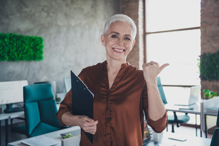 Photo of senior businesswoman point empty space hold papers wear brown shirt executive business leader modern office interior workspaceの写真素材