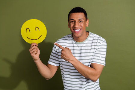 Photo of attractive funny guy wear striped t-shirt pointing smiling face emtpy space isolated khaki color backgroundの写真素材