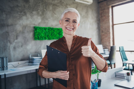 Photo of mature old female hold papers thumb up approve dressed brown shirt office professional workshop room interiorの写真素材