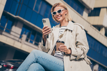 Photo of attractive young cheerful blonde lady hold telephone coffee paper cup wear trench coat walking outside outdoorsの写真素材