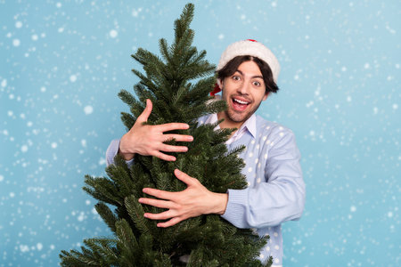 Photo of cheerful amazed crazy happy man hug christmas tree smile isolated on pastel blue color backgroundの写真素材