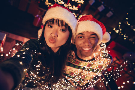 Photo of two young peaceful partners take selfie smile kiss eye wink festive christmas eve illumination apartment indoorsの写真素材