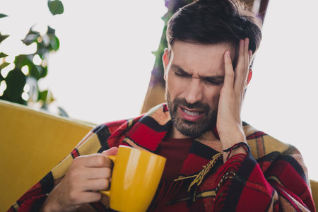 Young man with headache, dressed in a cozy plaid scarf, holding a warm beverage, relaxing at home.の写真素材
