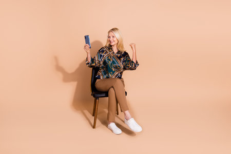 Photo of retired business woman sitting chair raised fist up victory in online casino on smartphone isolated over beige color backgroundの写真素材