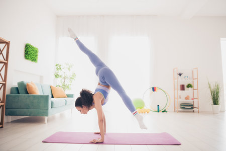 Young woman practicing handstand on yoga mat in bright home interior, showcasing strength and flexibility in trendy sportswearの写真素材