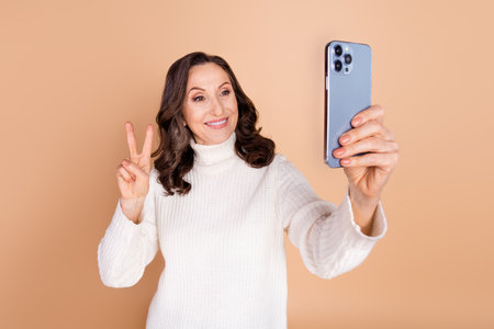 Photo of attractive adorable senior woman wear white stylish clothes hold gadget v-sign isolated on beige color backgroundの写真素材