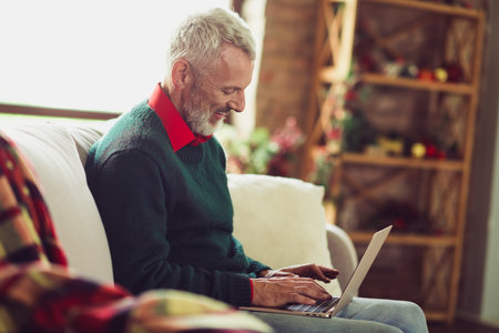 Photo of nice aged man sit couch use laptop wear sweater christmas festive time apartment indoorsの写真素材