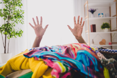 Young woman raising hands in colorful clothing pile during daylight inside homeの写真素材