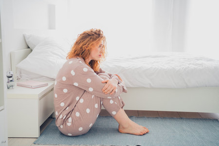 Charming young woman with red hair sitting in pajamas by the bed looking peaceful in a bright bedroomの写真素材