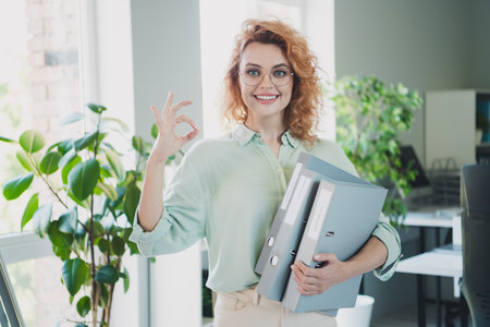 Photo of successful young business lady hold folders show okey symbol wear shirt modern interior office indoorsの写真素材