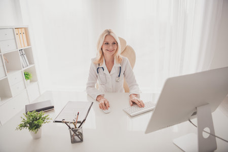 Confident senior female doctor working on computer in modern hospital officeの写真素材