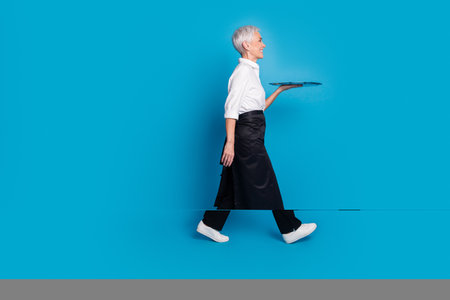Full size photo of retired female walk profile carry tray barista small coffee shop owner wear waitress uniform isolated on blue backgroundの写真素材