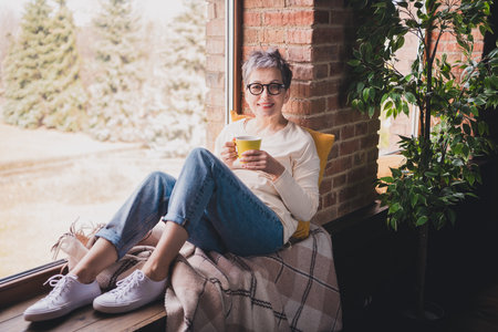 Charming mature woman with grey hair and glasses, sitting casually by a bright window with a warm smile, enjoying a relaxing momentの写真素材