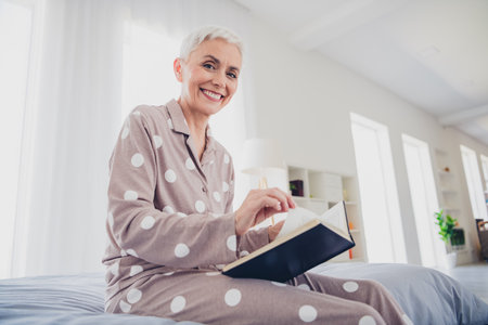 Elderly woman with grey hair enjoying morning reading in cozy bedroomの写真素材