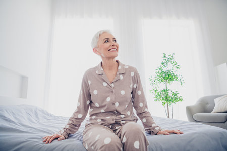 Charming elderly woman with grey hair enjoying morning light in cozy bedroomの写真素材