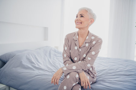 Charming elderly woman with grey hair smiling joyfully in stylish pajamas, sitting on a cozy bed in a bright bedroomの写真素材