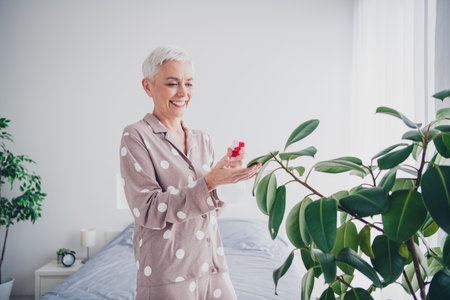 Elderly woman enjoying morning in pajamas with vibrant plants in bright bedroomの写真素材