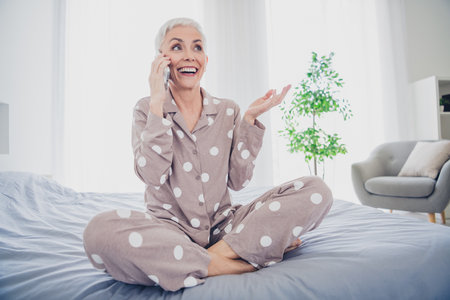 Elderly woman in polka dot sleepwear enjoying a morning chat, sitting on a bed in bright natural light.の写真素材