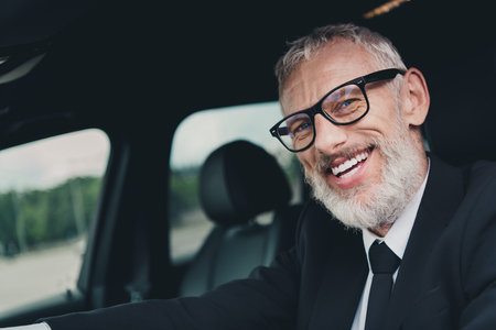 Smiling senior businessman in suit sits in car on a city street, exuding confidence and sophisticationの写真素材