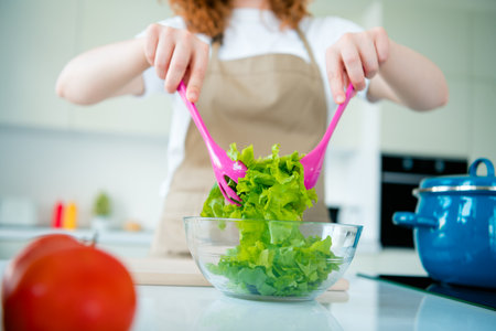Young woman in apron preparing fresh salad with pink utensils in bright kitchenの写真素材