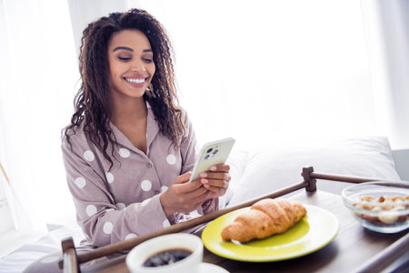 Young woman enjoying a relaxing morning in bed, checking her phone with a smile, wearing cozy polka-dot sleepwearの写真素材