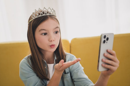 Adorable young girl with blond hair takes a selfie indoors, wearing a denim shirt and crown, exuding charm and happinessの写真素材