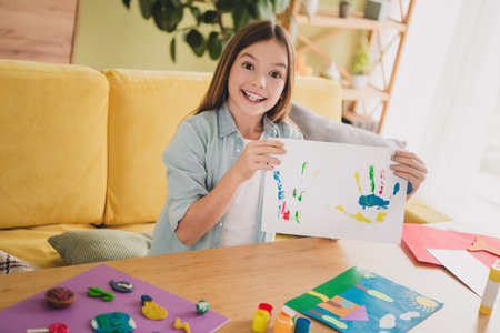 Adorable girl showcasing her creative artwork in a cozy living room with colorful handprints and art supplies on the tableの写真素材