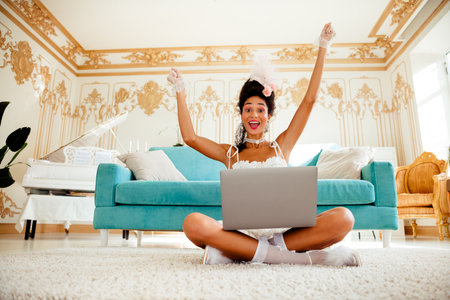 Vivacious woman in elegant baroque-inspired room embraces technology, celebrating success while sitting comfortably in luxurious interiorの写真素材