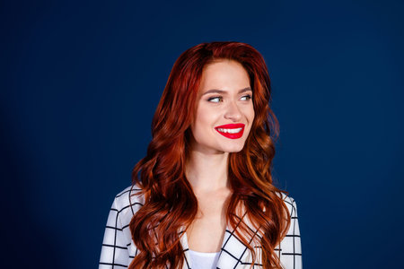 Young woman with red hair and bright smile wearing a trendy checkered suit, exudes elegance and charm against a dark blue backgroundの写真素材