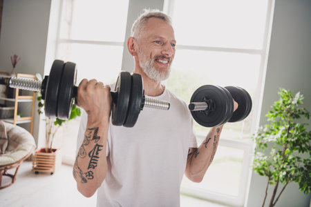 Elderly man enjoying his healthy lifestyle by lifting weights at home, showcasing fitness and strength in a bright indoorの写真素材