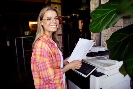 Photo of attractive young blonde woman use photocopier wear plaid shirt modern dark business center coworking interior indoors workspaceの写真素材