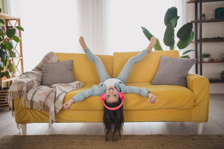 Charming young girl in casual denim playfully upside down on the sofa with pink headphones, enjoying a relaxing weekend at home.の写真素材