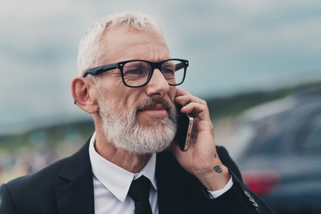 Mature businessman in suit talking on phone outdoors near automobile, showcasing travel and work...