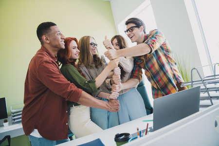 Photo of group young corporate workers agree pile stack arms thumb up finger workstation office business center indoorsの写真素材