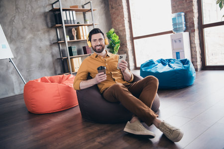 Full body photo of man sit beanbag coffee gadget listen music wear yellow shirt formalwear modern office room interior indoors workspaceの写真素材