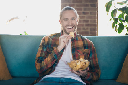 Young man with a beard happily enjoying snacks at home in a relaxed and stylish loft environmentの写真素材