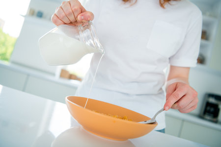 Young woman in casual style pouring milk into cereal for breakfast in bright kitchen
