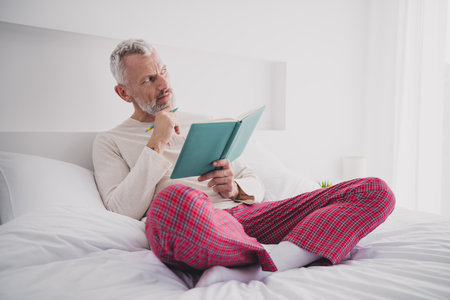 Mature man in cozy pajamas enjoying a thoughtful morning in bed while reading a bookの写真素材