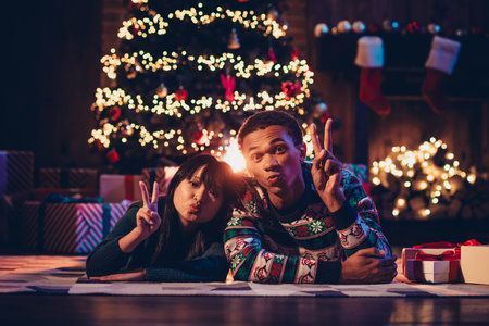 Full length photo of happy young couple lying floor v-sign air kiss dressed christmas garment tradition atmosphere decor house indoors roomの写真素材