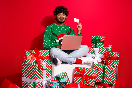 Full length photo of young guy sit pile gifts netbook credit card wear green christmas sweater celebrate winter holidays on red backgroundの写真素材