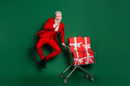 Full length portrait of stylish santa jump presents pushcart red christmas suit isolated on green color backgroundの写真素材