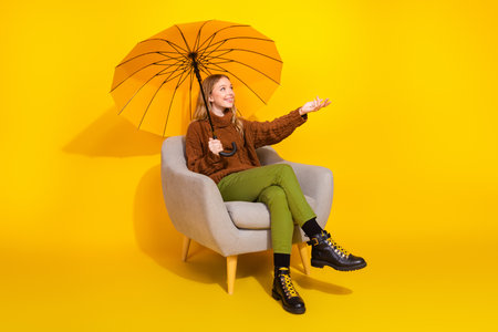 Full size photo of pretty young woman sit chair umbrella arm catch empty space raindrop isolated on yellow color backgroundの写真素材